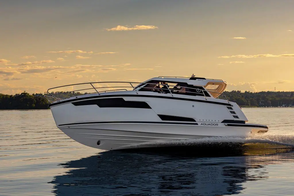 A sleek white boat named Aquador glides over calm water at sunset, with trees and clouds in the background.